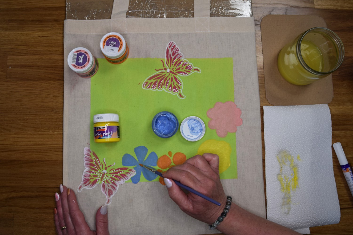 Žena maľuje na plátennú tašku kvety rôznymi odtieňmi farieb.